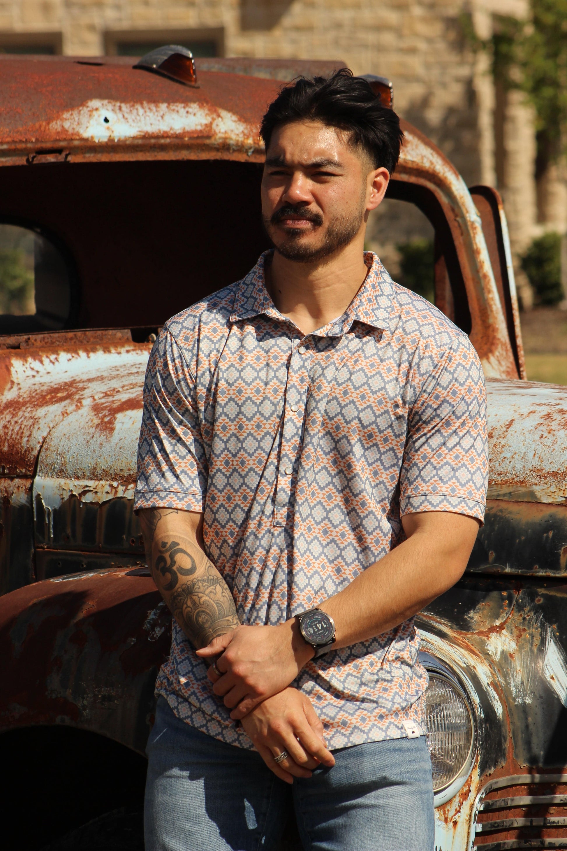 A man stands in front of a vintage rusted truck, modeling the HowdyHo Aztec Twilight Blend shirt. The short-sleeved shirt boasts a complex Aztec pattern in shades of blue and orange, offering a modern twist on classic southwestern motifs. His casual stance, with arms crossed, along with the rugged outdoor backdrop, highlights the shirt's adventurous spirit and durability.