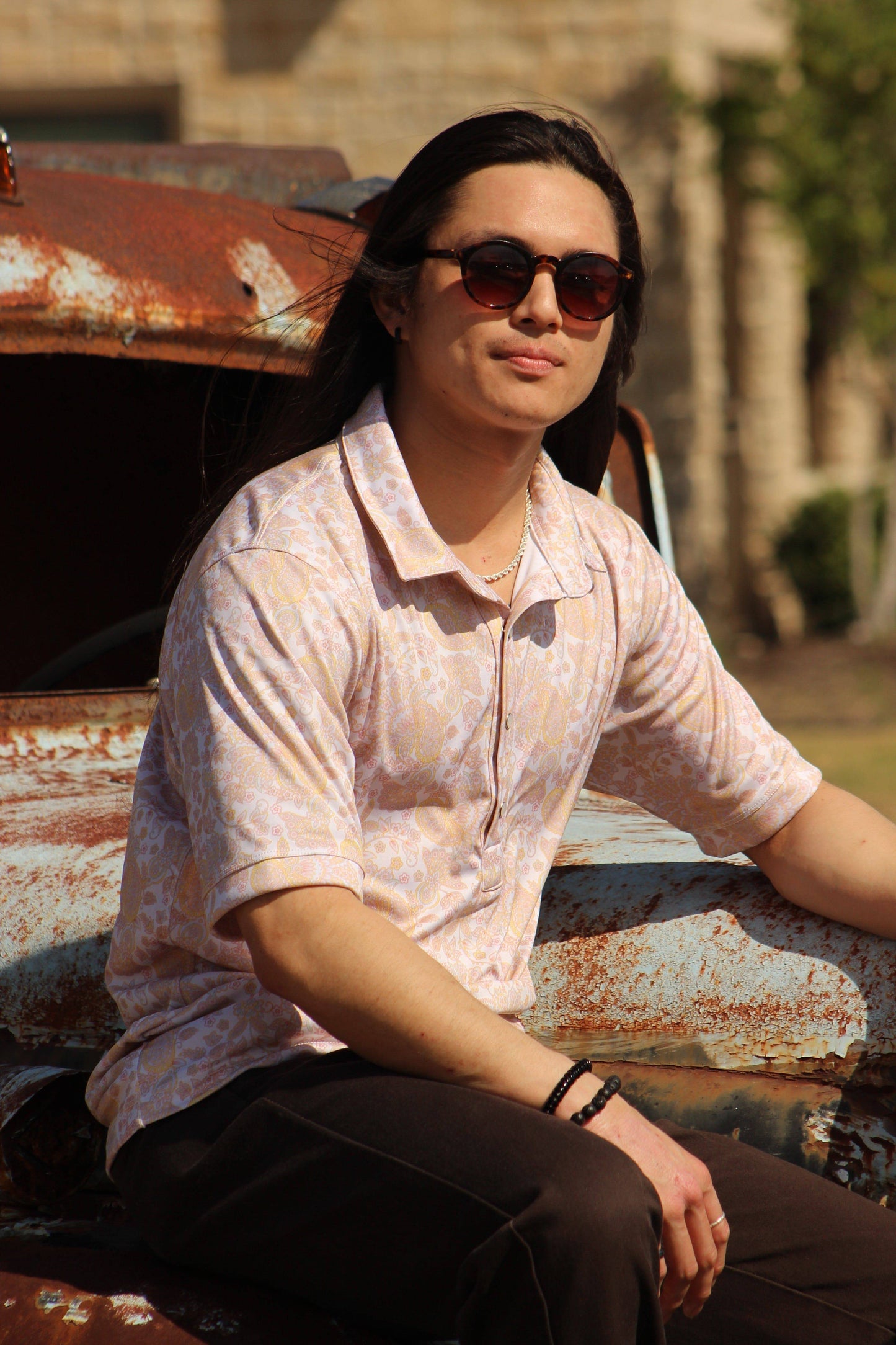 A person exudes a relaxed yet stylish vibe in a HowdyHo Sunshine Paisley shirt, its subtle paisley pattern in warm peach tones offering a fresh take on a classic design. The shirt's fit is complemented by dark trousers and cool sunglasses, perfect for a casual outing. They are seated on a weathered, rusted metal surface, adding an edgy contrast to the soft shirt pattern.