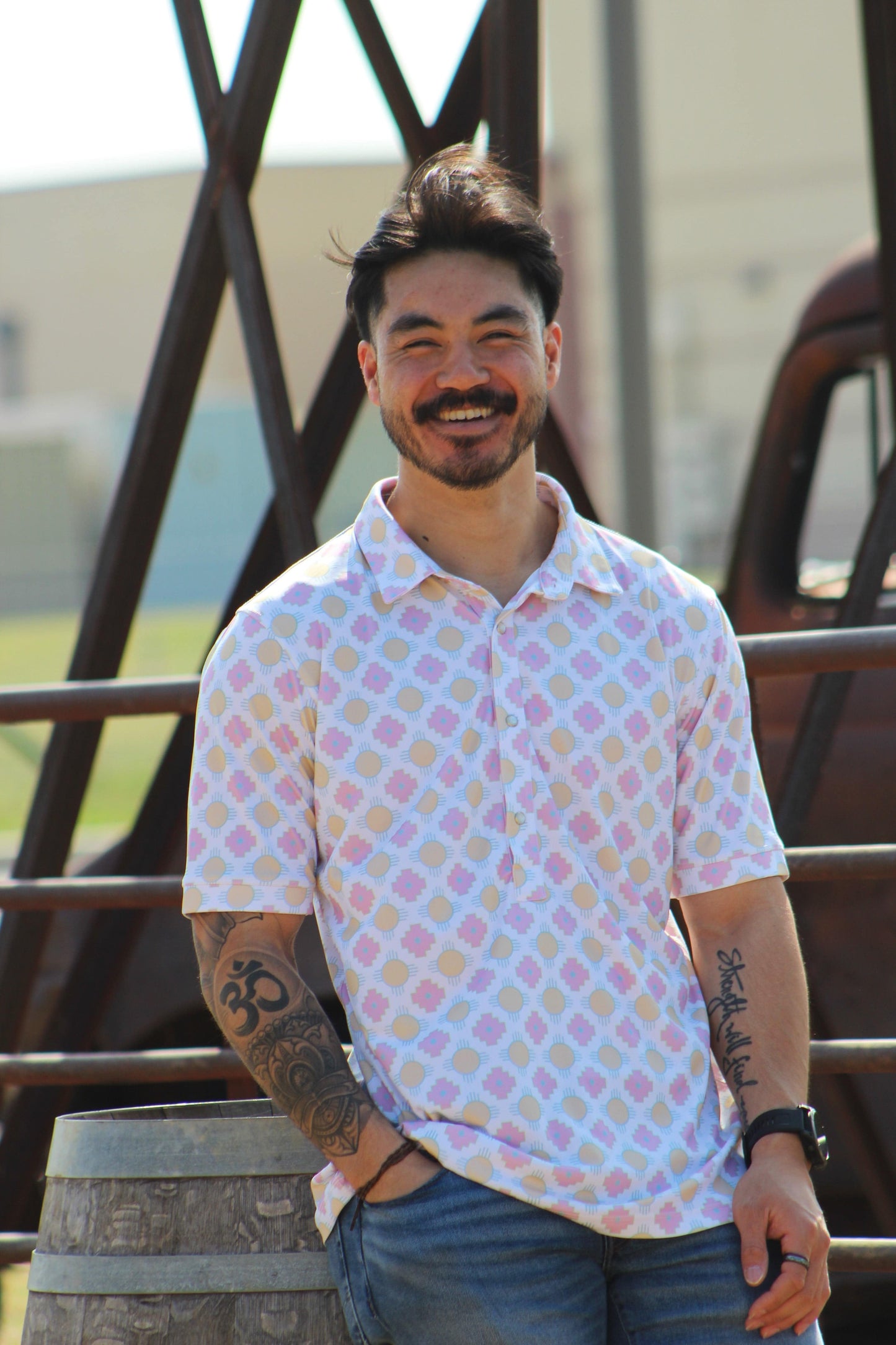 A man with a warm smile is casually leaning on a wooden barrel, wearing the Sunset Crossroads shirt from HowdyHo. The shirt boasts a soft pattern of yellow suns and pink crosses on a light background, creating a fun and fresh look. His relaxed pose, paired with jeans and a stylish watch, captures the essence of HowdyHo's blend of comfort and trendy design.