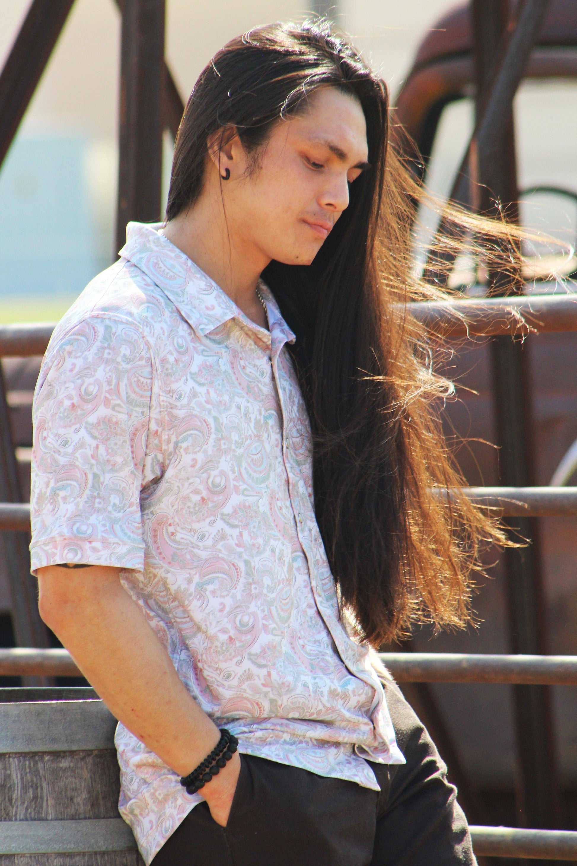 A close-up view of a man wearing the Green Love Paisley shirt, capturing the intricate paisley print and subtle pastel hues. The shirt features a relaxed fit and a pointed collar, styled with the sleeves casually rolled up. His long, wind-touched hair adds a sense of movement to the image, emphasizing the lightweight fabric of the shirt.
