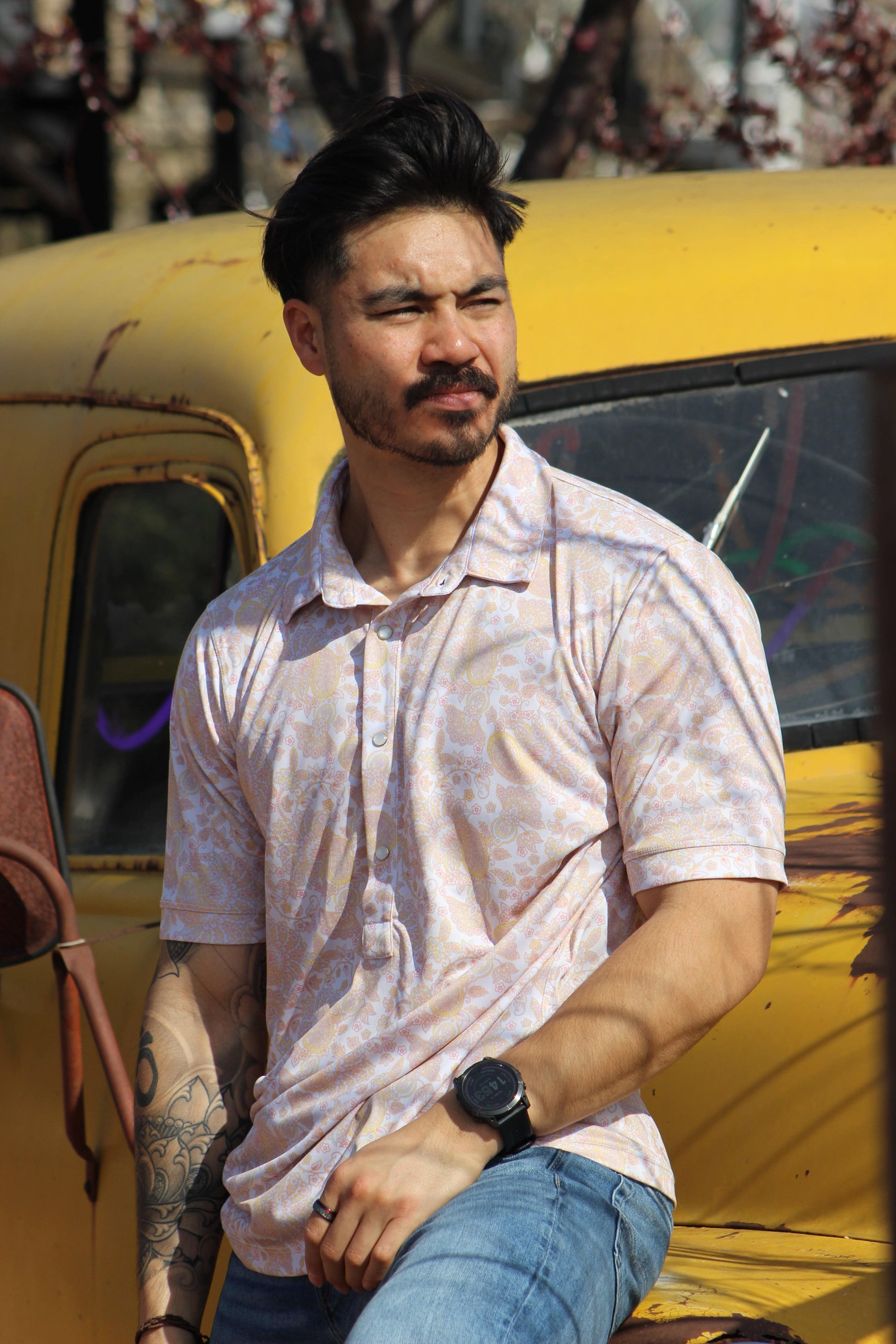 A rugged man with a stylish beard and tattooed arm models the HowdyHo Sunshine Paisley shirt. The shirt's light paisley pattern adds a touch of understated elegance to his laid-back look. He's casually leaning against a vintage yellow truck, with a black watch on his wrist, embodying a blend of masculinity and sophistication.