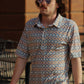 A man walks confidently in an urban setting, wearing the Aztec Twilight Blend shirt from HowdyHo. This stylish piece features a unique blue and orange Aztec pattern on a crisp, button-down shirt with a pointed collar. The shirt's contemporary design is complemented by his cool round sunglasses, adding a modern twist to a classic look.