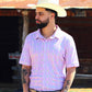 A man stands poised outdoors, sporting the Rose Quetzal shirt from HowdyHo, distinguished by its pink vertical stripes and diamond-shaped motifs. The shirt is styled with a classic collar and the sleeves are neatly cuffed, paired with a stylish beige cowboy hat, dark blue jeans, and a black leather belt. His confident stance and the rustic barn in the background create an authentic Western vibe.