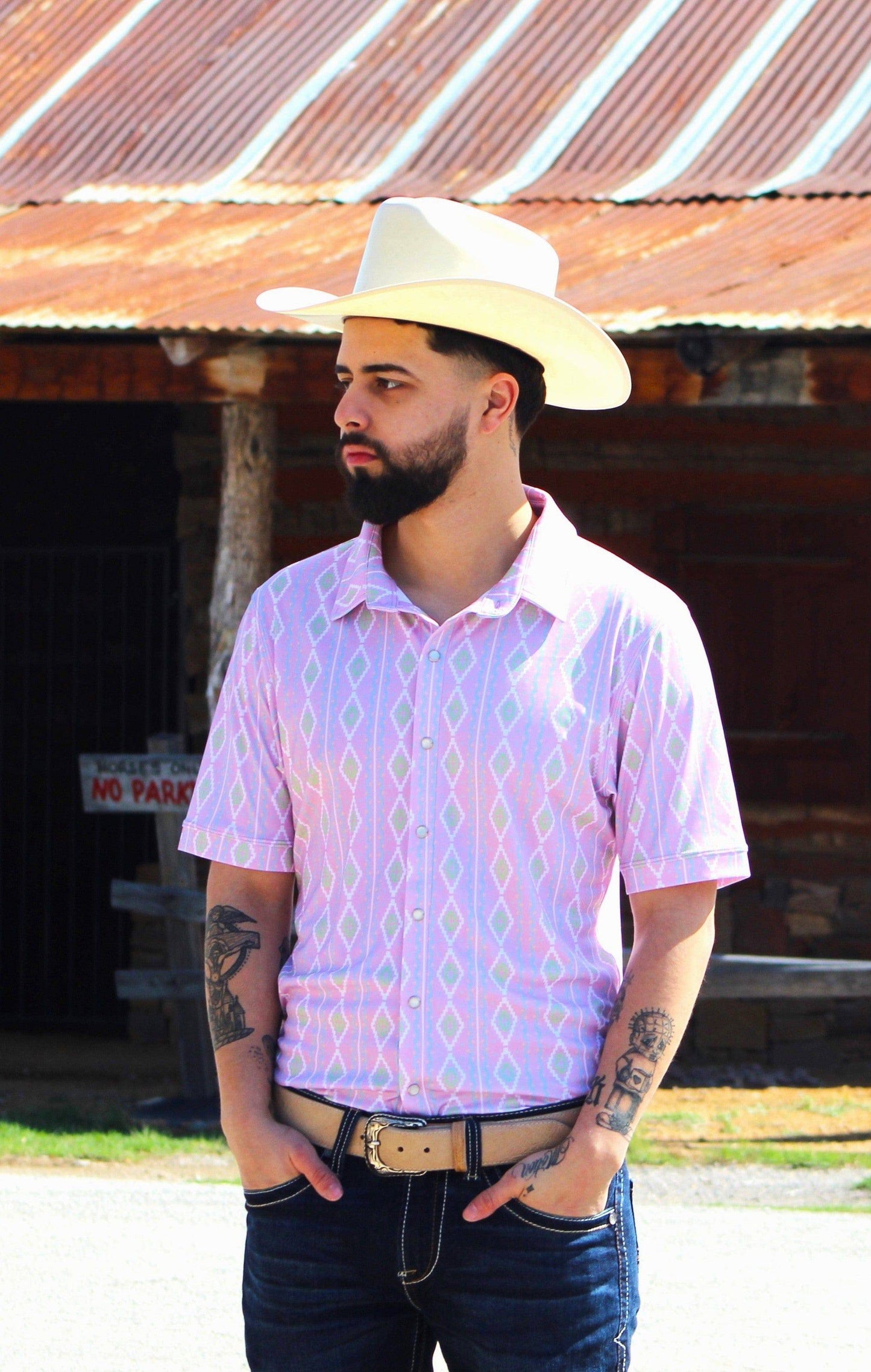 A man stands poised outdoors, sporting the Rose Quetzal shirt from HowdyHo, distinguished by its pink vertical stripes and diamond-shaped motifs. The shirt is styled with a classic collar and the sleeves are neatly cuffed, paired with a stylish beige cowboy hat, dark blue jeans, and a black leather belt. His confident stance and the rustic barn in the background create an authentic Western vibe.