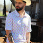 A man projects a relaxed yet confident demeanor in the Sunset Crossroads shirt by HowdyHo, featuring a playful pattern of pink and yellow suns on a light background. His classic cowboy hat and a well-fitted pair of jeans complete the ensemble, offering a modern twist on traditional Western style. The industrial backdrop adds an edgy contrast to the shirt's soft design.