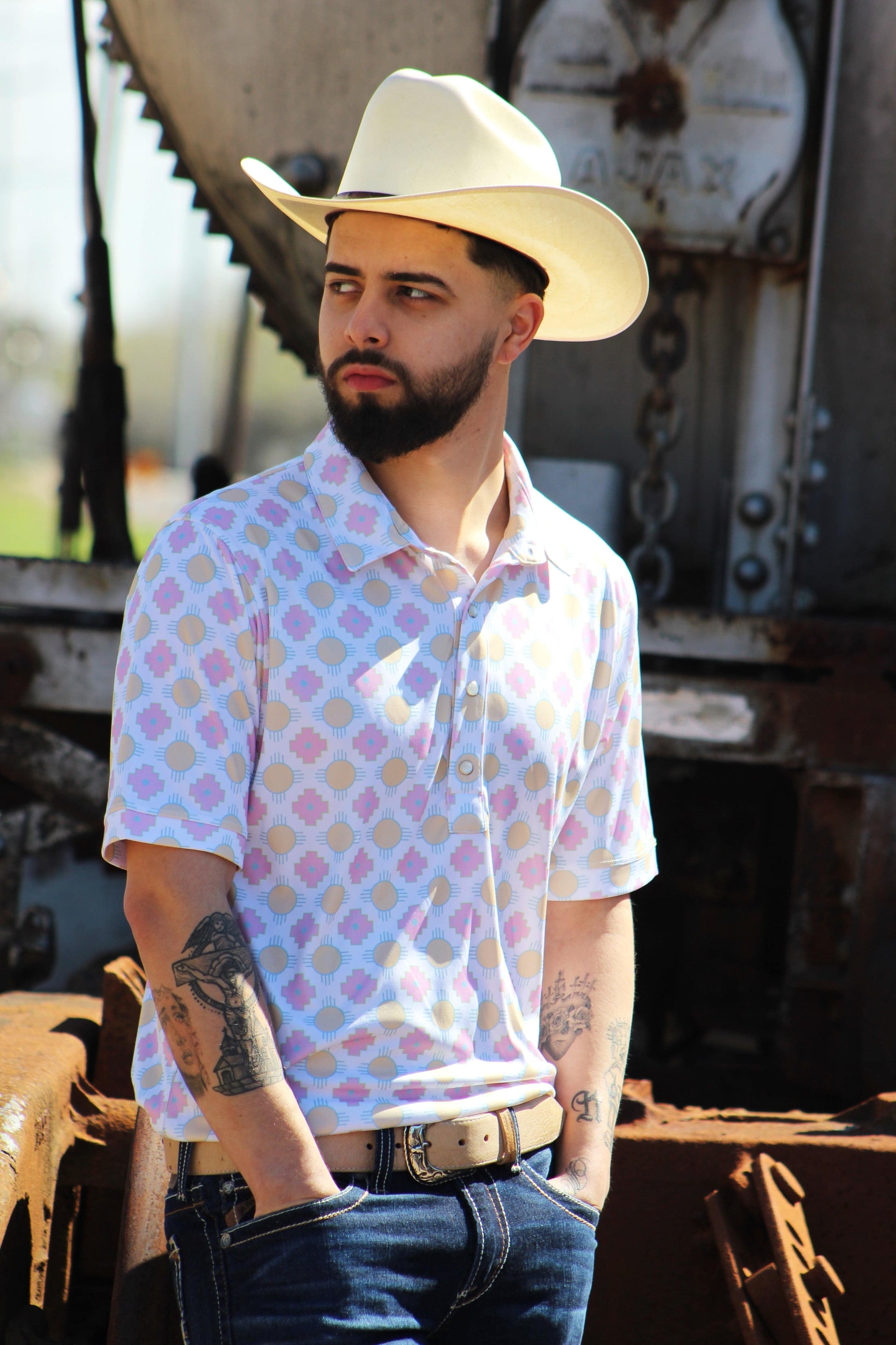 A man projects a relaxed yet confident demeanor in the Sunset Crossroads shirt by HowdyHo, featuring a playful pattern of pink and yellow suns on a light background. His classic cowboy hat and a well-fitted pair of jeans complete the ensemble, offering a modern twist on traditional Western style. The industrial backdrop adds an edgy contrast to the shirt's soft design.