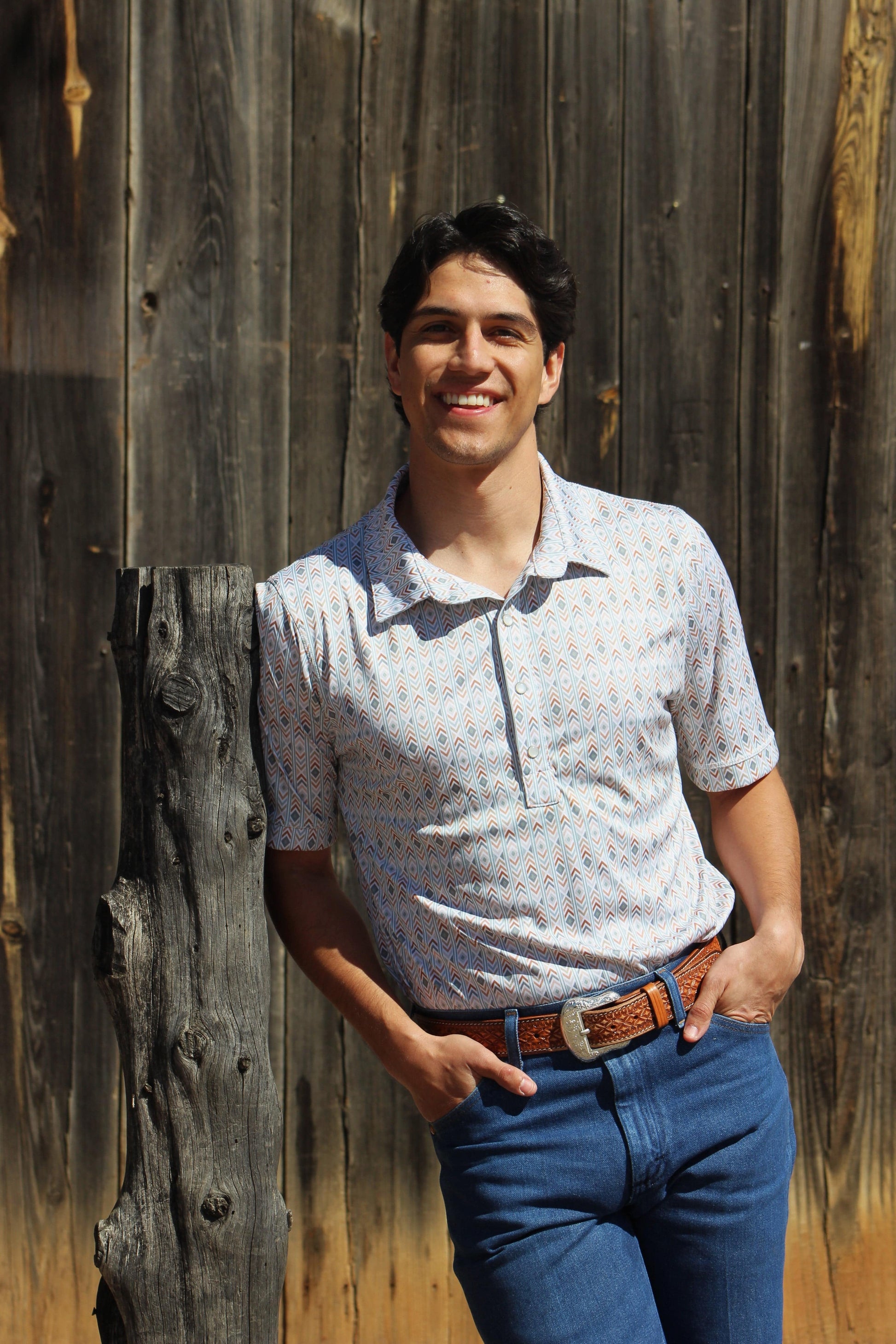 A happy young man poses confidently in HowdyHo's Aztec Fusion short-sleeved shirt, paired with blue jeans and a brown leather belt. The shirt's unique geometric print in soft hues complements the rustic wooden backdrop, capturing the essence of a casual, modern style.